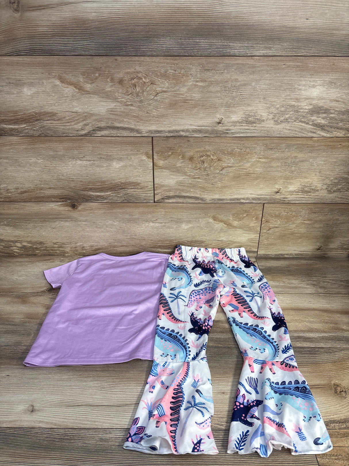 Flat lay of purple short-sleeve shirt and white flare pants with pastel dinosaur print and ruffled cuffs on wood floor