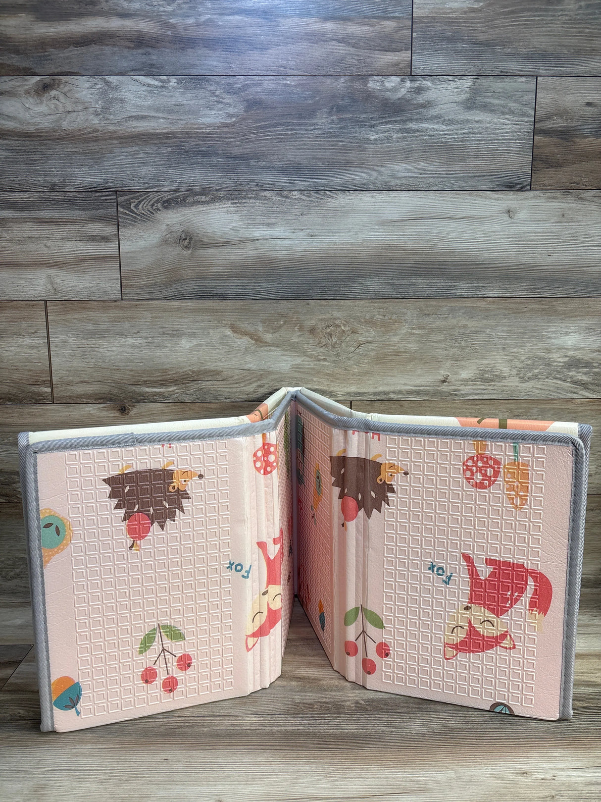 Foldable pink baby play mat with cartoon animals and fruit on wood floor