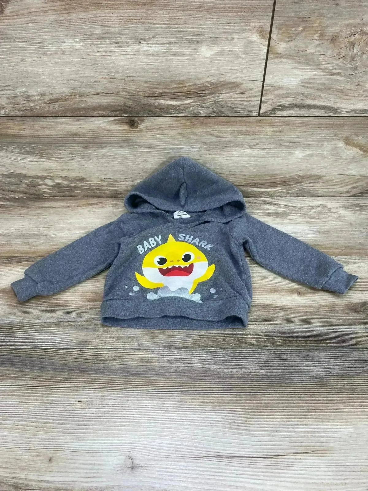 Front view of Pinkfong Baby Shark grey hoodie, size 12 months, showing yellow Baby Shark graphic and attached hood on wood background