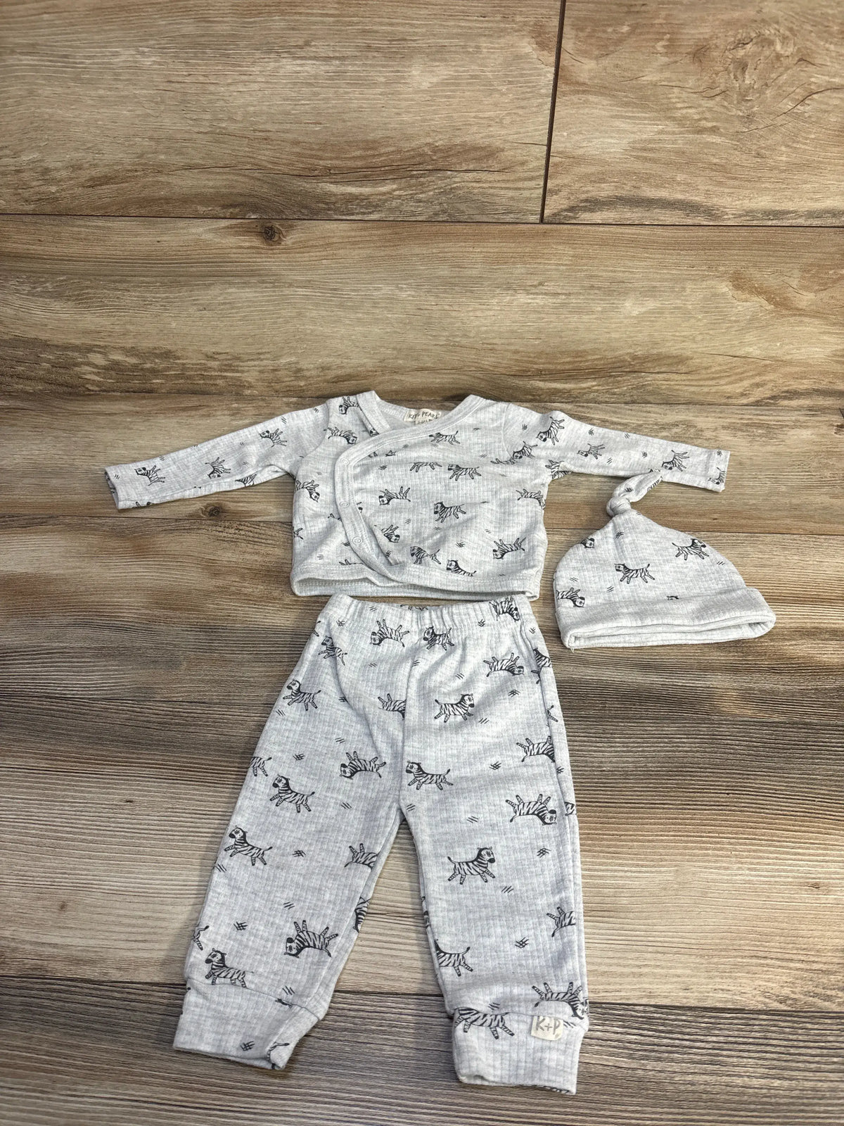 Flat lay of Kit + Pearl three-piece zebra print set in grey, showcasing shirt, pants, and hat on a wood floor