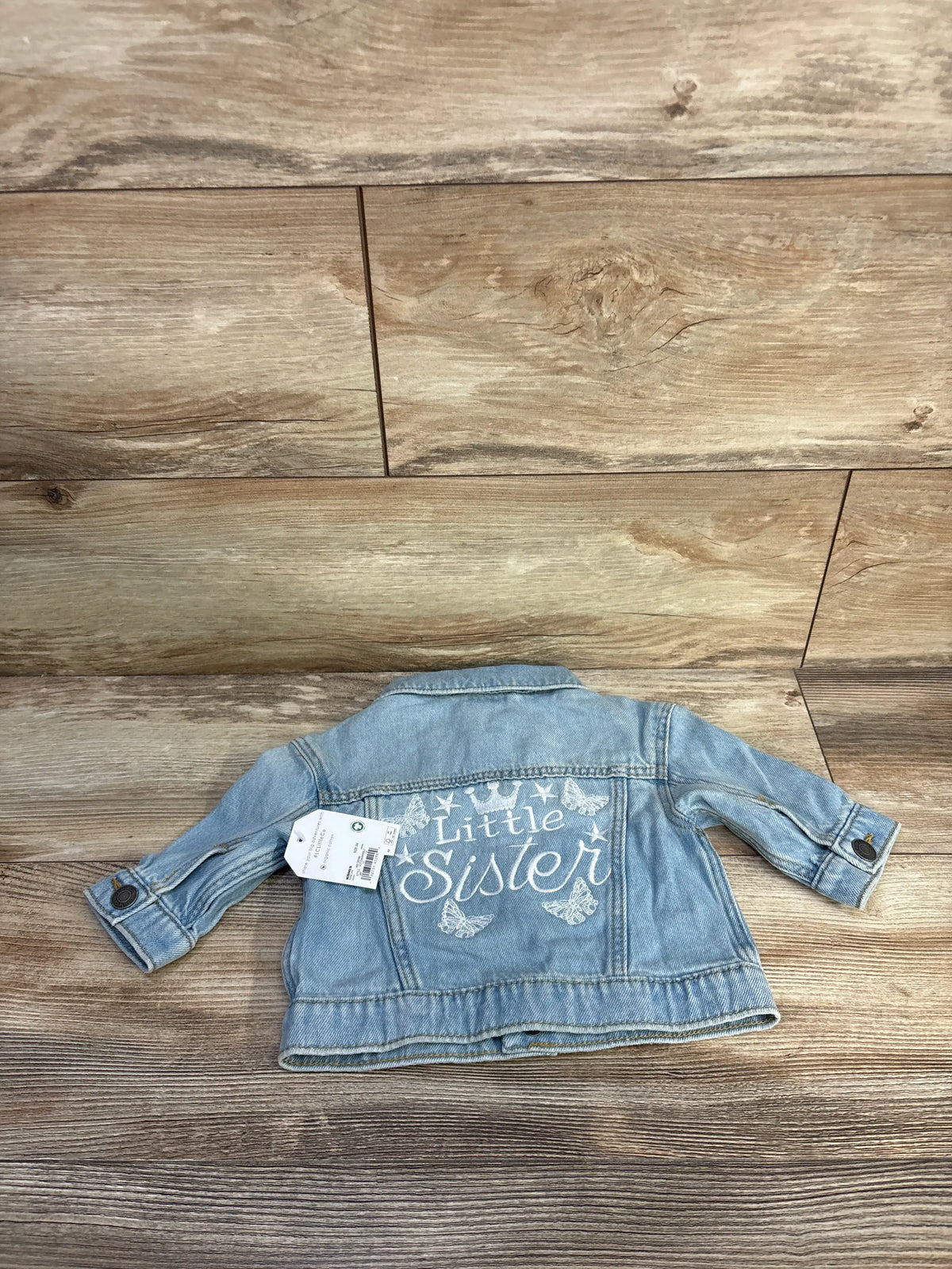 Back view of Little Co. blue denim jacket showcasing white Little Sister embroidery with wood-look floor background