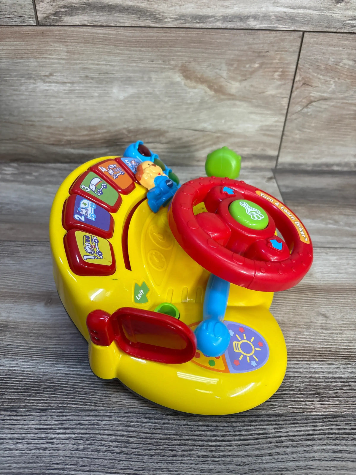 Angled view of Vtech Turn & Learn Driver showcasing steering wheel, colorful buttons, signal lever, and mirror on a wood-grain background