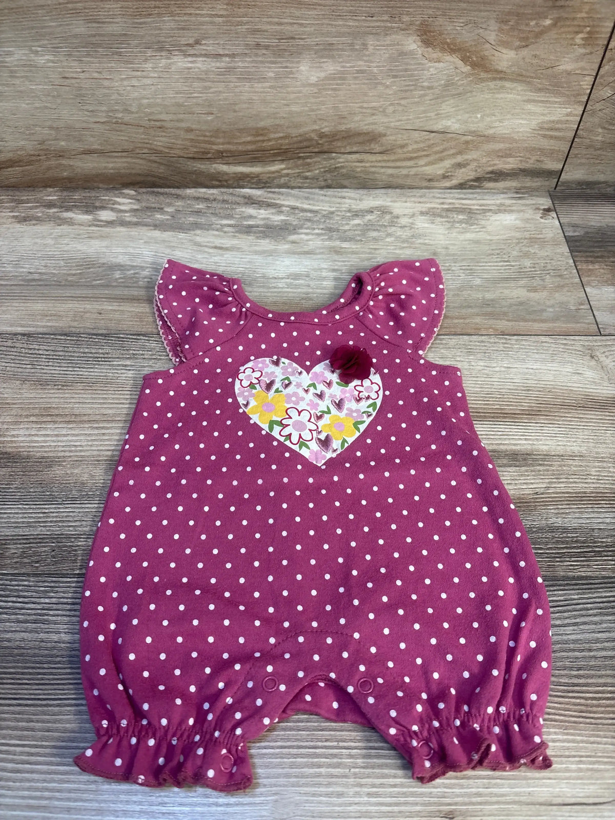 Top view of mauve polka dot baby shortie romper showcasing ruffle sleeves, floral heart applique and snap crotch on wood background