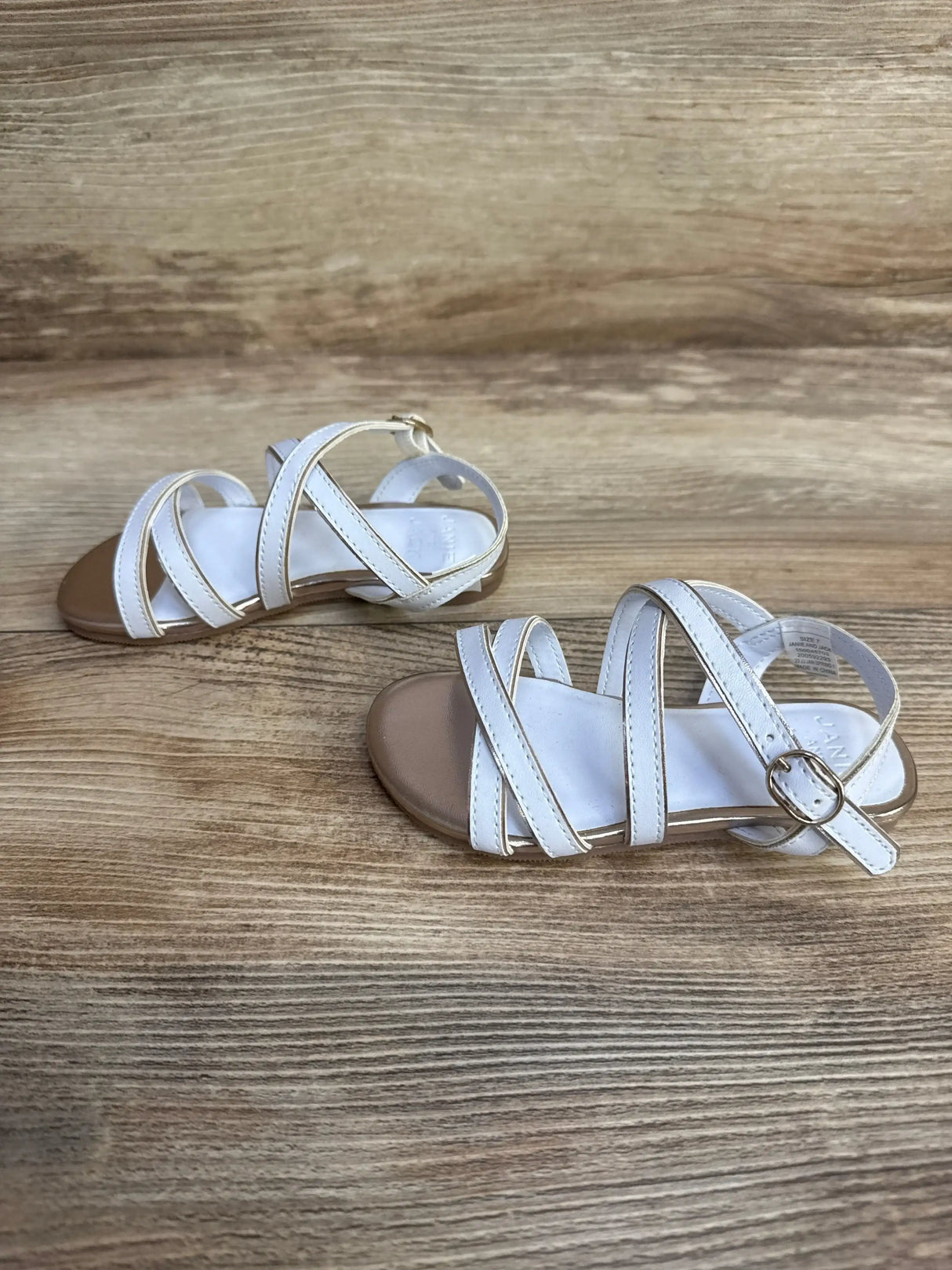Three-quarter top view of toddler white sandal showcasing gold metallic trim on crossed straps and ankle buckle with wood plank background