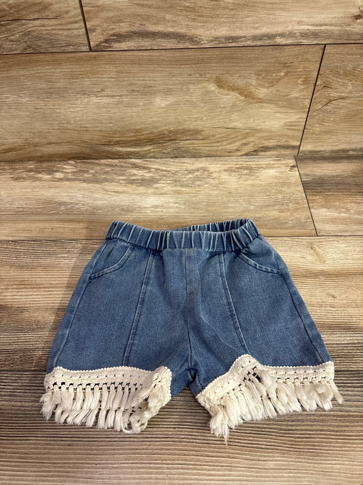 Blue pull-on denim shorts for toddlers with white tassel trim hem on a wood background