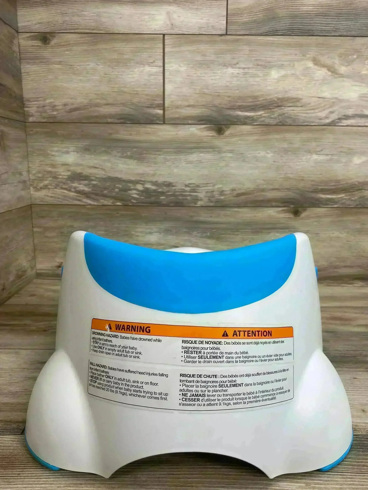 Rear view of Munchkin Clean Cradle baby bathtub showcasing blue contoured headrest and warning label on wood plank background
