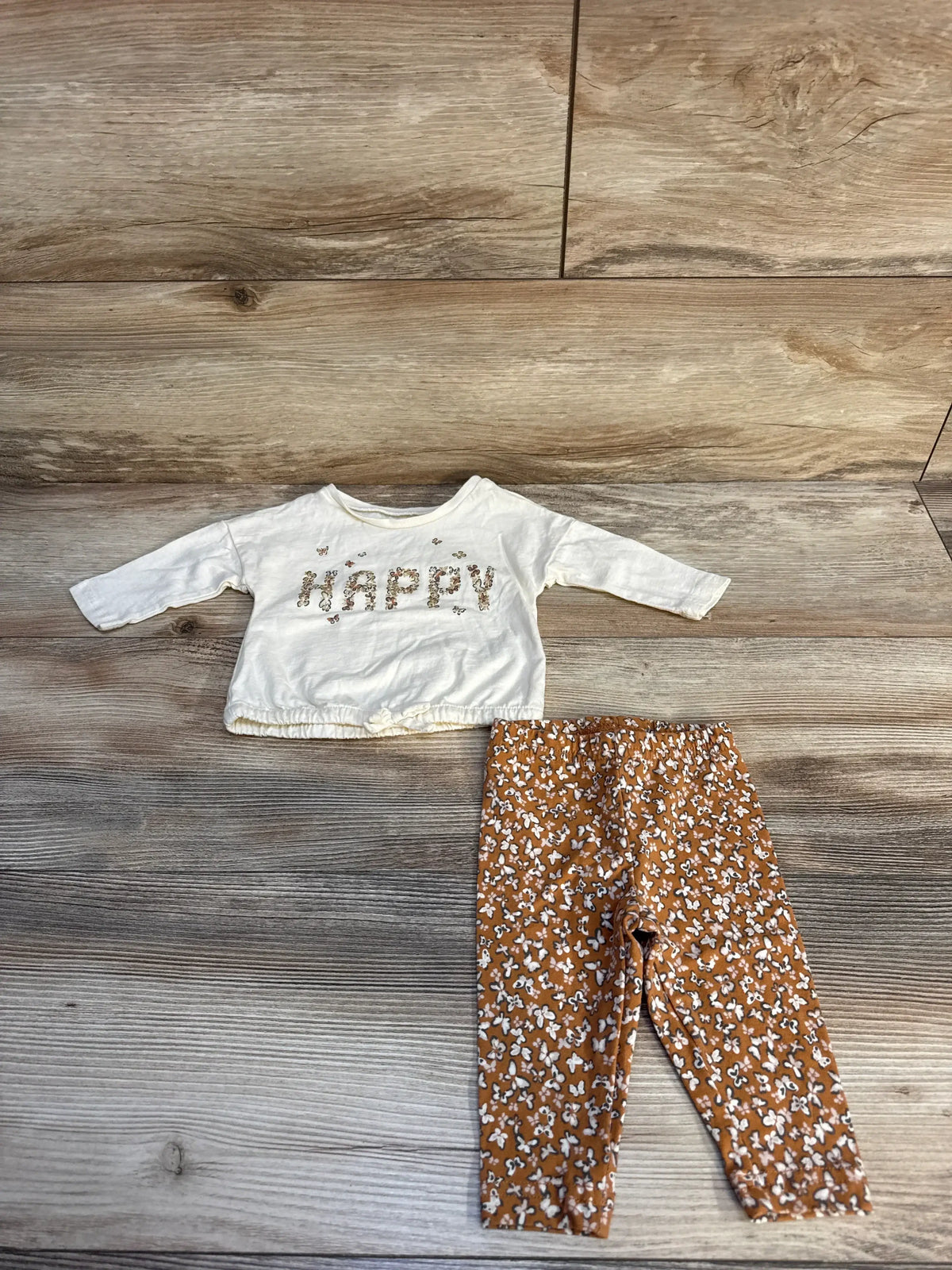 Flat lay of Carter’s Happy Butterfly 2-piece set (3M) showing ivory long-sleeve top with ‘HAPPY’ floral letters and gathered hem bow, and rust-brown floral leggings on wood floor