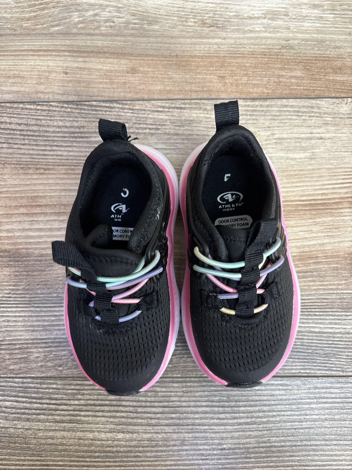 Top view of black kids' athletic shoes with pink soles and multicolor elastic laces on wooden floor