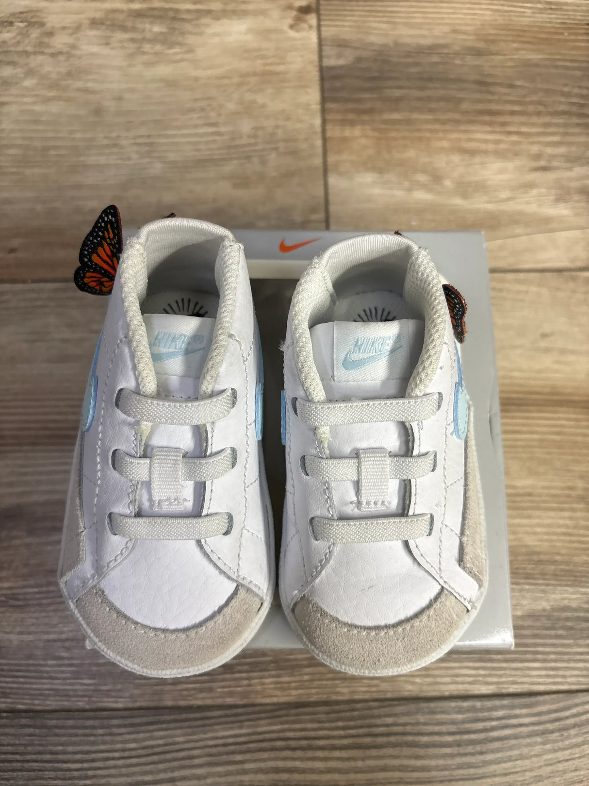 Pair of white Nike baby shoes with light blue logo and beige toe caps on wooden floor