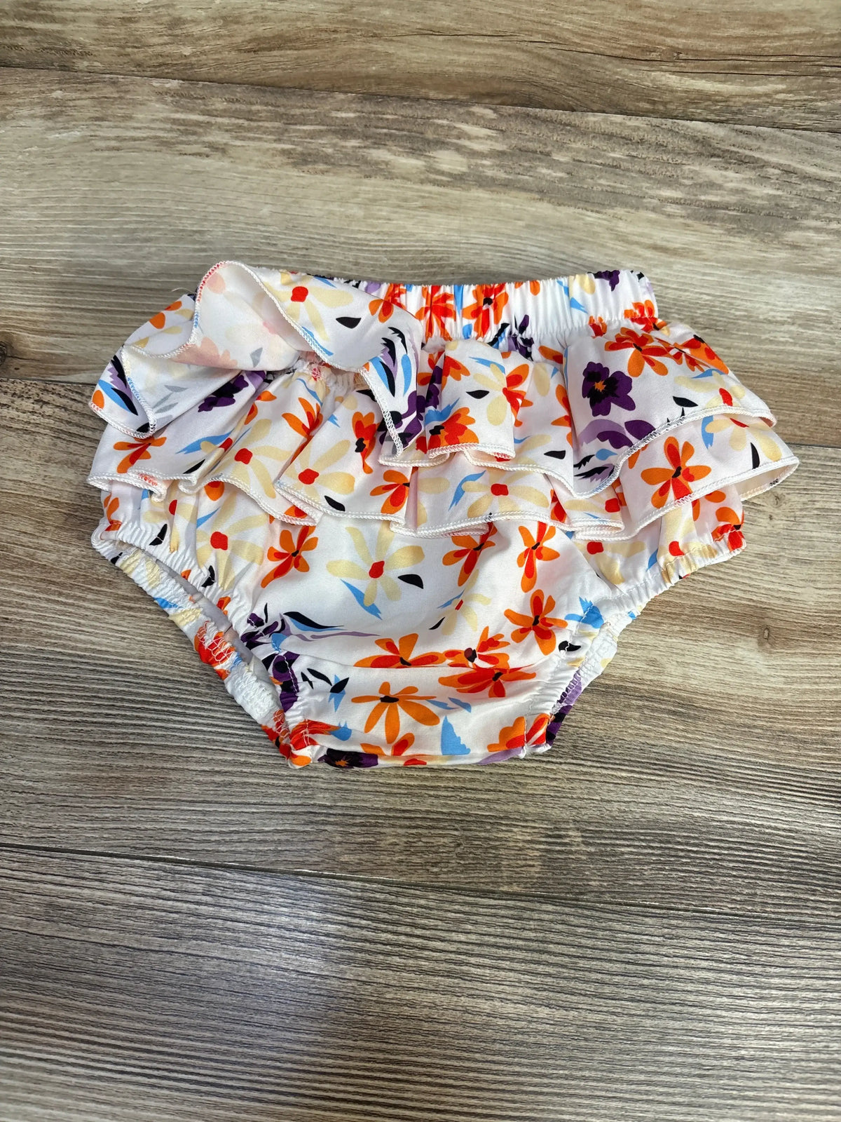 Flat lay of white floral ruffle baby bloomers showcasing double ruffle trim, elastic waist and leg openings on wooden floor background