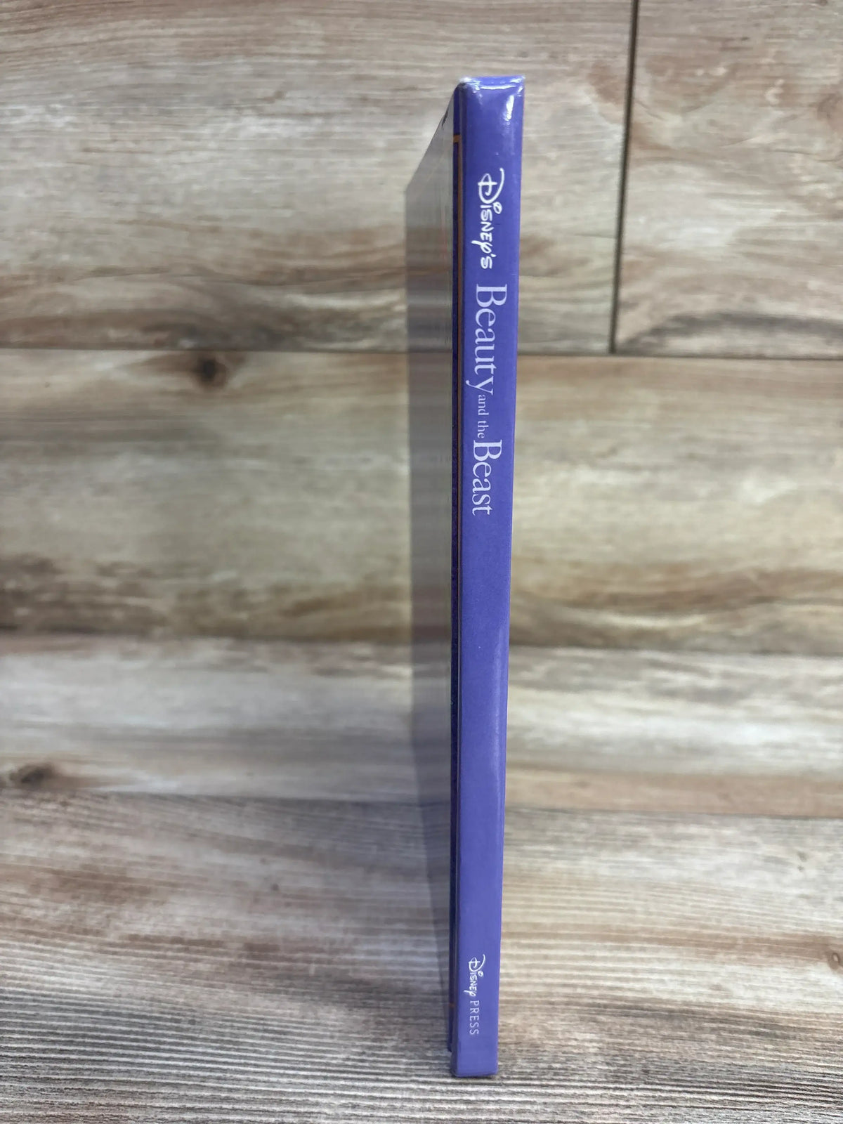 Side view of Disney Beauty and the Beast hardcover book showing purple spine with vertical title and Disney Press logo, shrink-wrapped on wood background