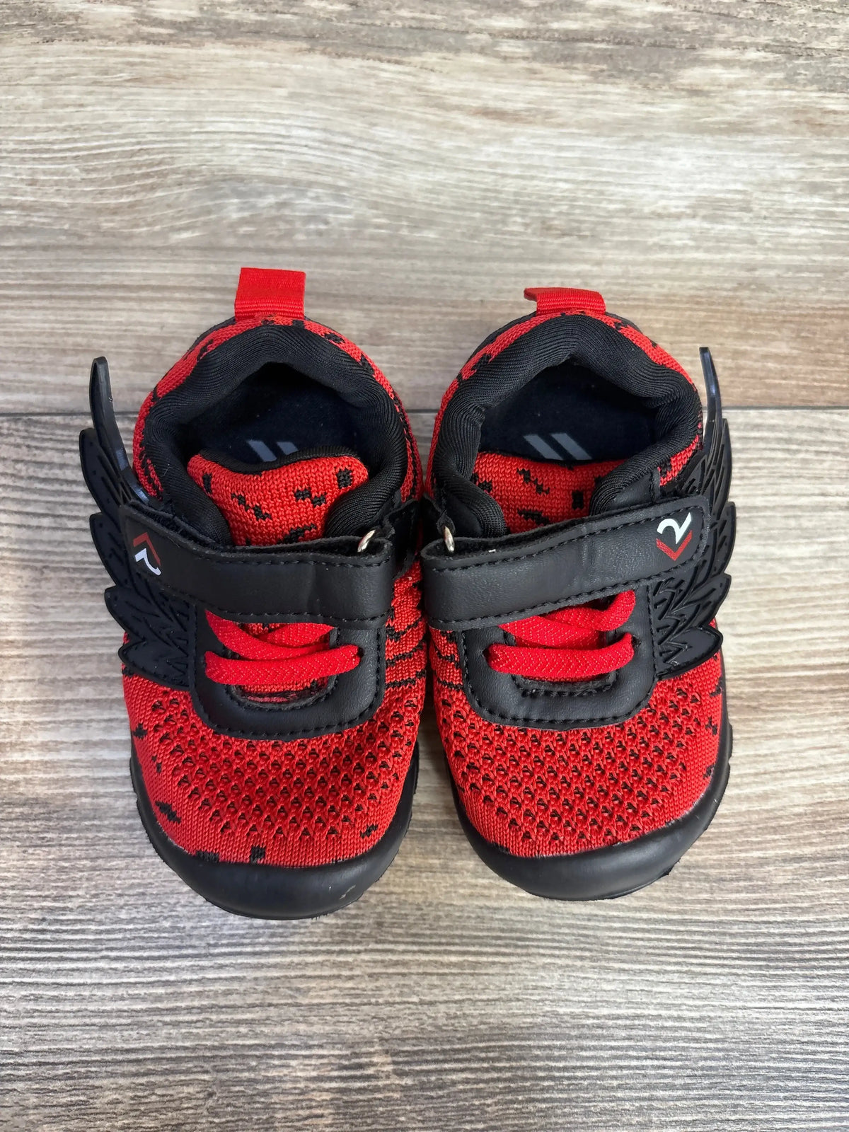 Top-down view of boy's red and black wing sneakers size 3.5C with knitted mesh upper and black velcro wing strap on wood background