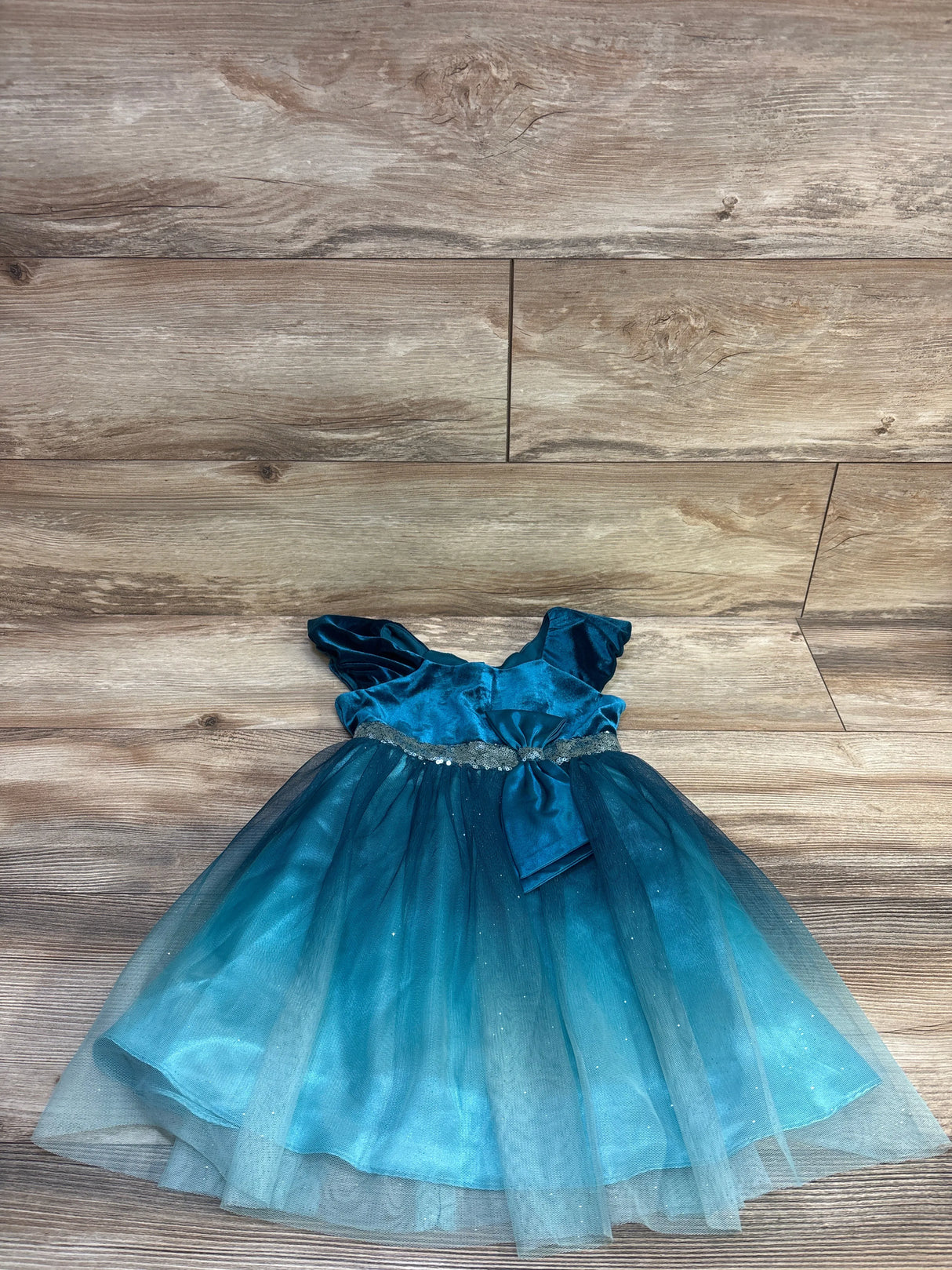 Flat lay of teal Jona Michelle velvet tulle toddler dress showcasing sequin-trimmed waist, decorative bow and layered glitter tulle on wood floor