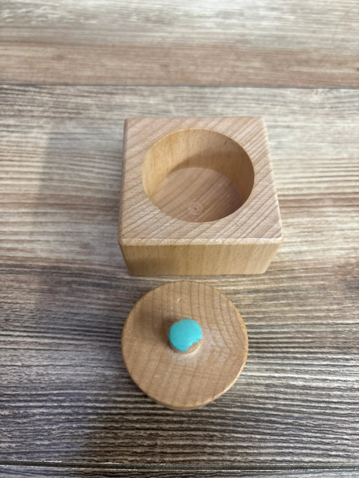 Top-down view of Lovevery Pincer Puzzle, wooden box with circular recess and removable lid with turquoise knob on wood table