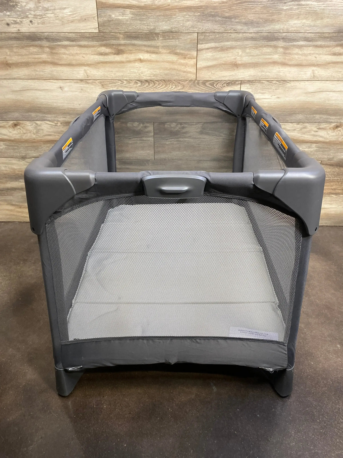 Front view of 4moms Breeze Go playard in grey showcasing mesh sides and mattress pad with wood-panel wall background