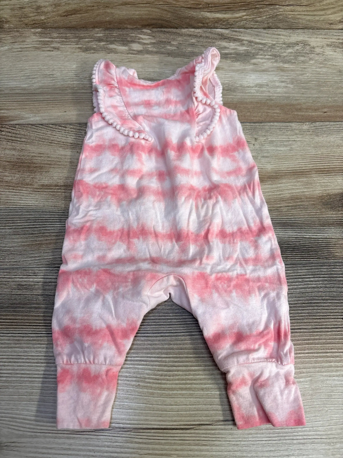 Top-down view of pink tie-dye baby romper (0-3M) showcasing ruffled pom-trim collar and cuffed ankles on wood background