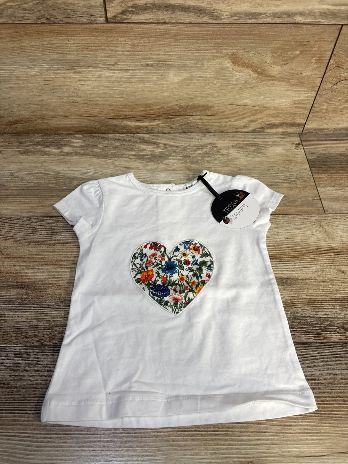 Front view of white Tessa James Birdie Tee showcasing floral heart appliqué and puff sleeves on a wood floor
