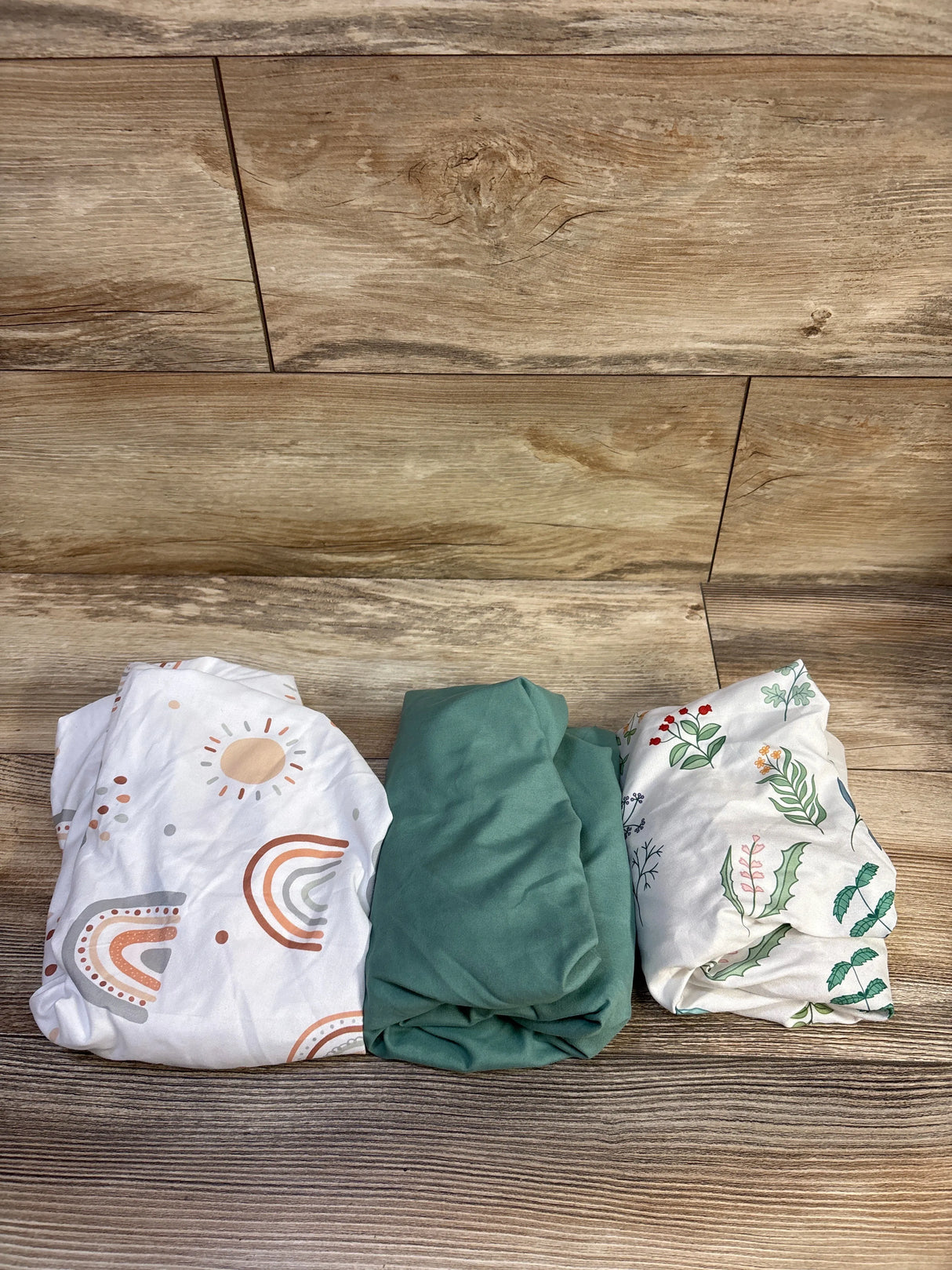 Top view of three fitted bassinet sheets: one solid green and two patterned (rainbow, botanical) on wood floor