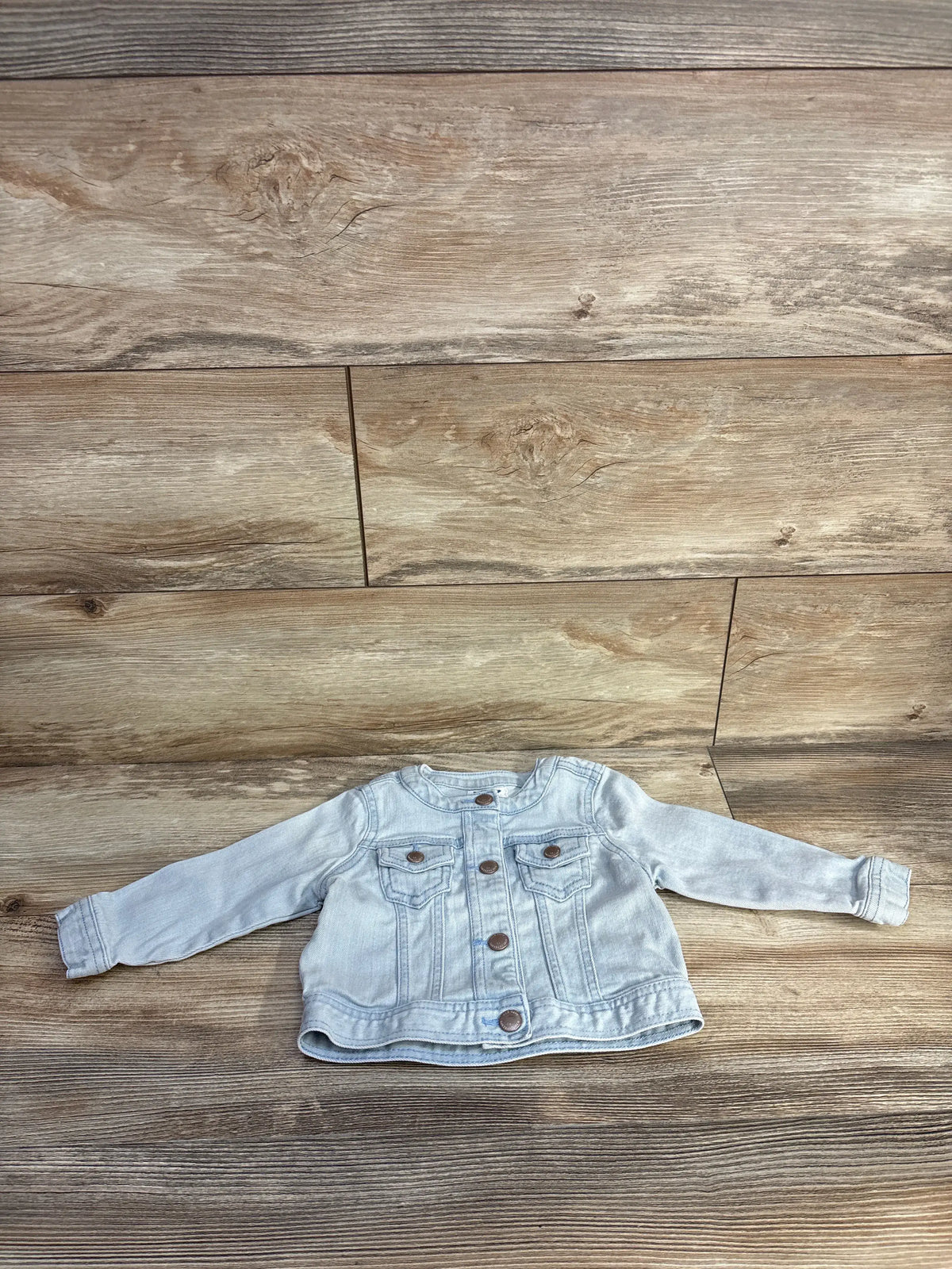 Front view of Old Navy blue denim jacket, size 12 to 18 months, showcasing button front and two chest pockets on wood background