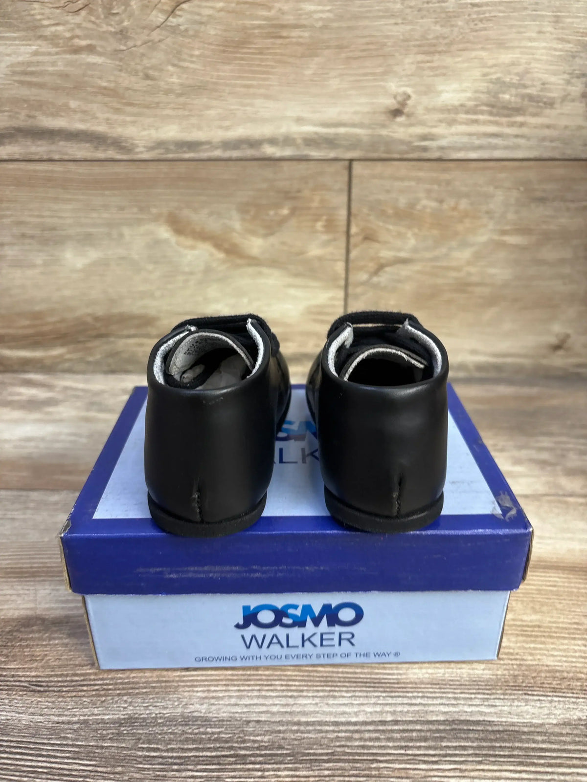 Rear view of black Josmo ankle baby shoes showing Velcro straps on blue Josmo Walker box with wood backdrop