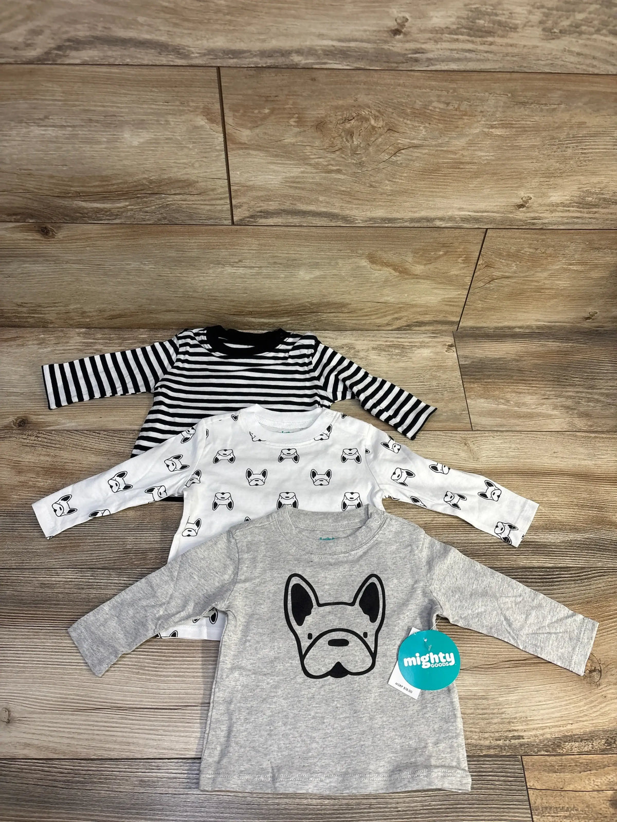 Flat lay of three Mighty Goods long sleeve dog print shirts in grey, white, and black stripes on a wood floor