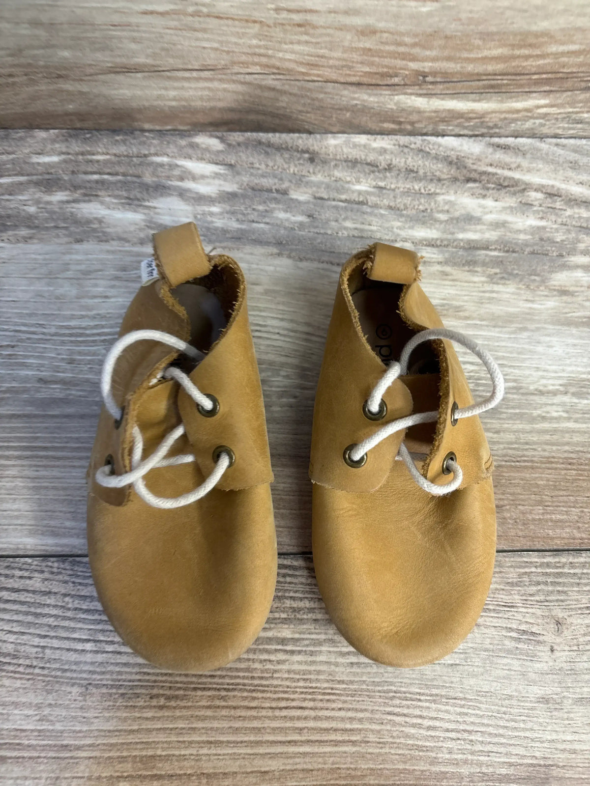 Tan leather baby shoes with white laces on wooden background, gently used kids footwear