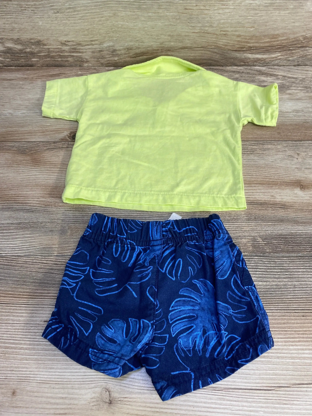 Flat lay of baby 2-piece set: green polo-style shirt and navy elastic-waist shorts with blue leaf print on wood background