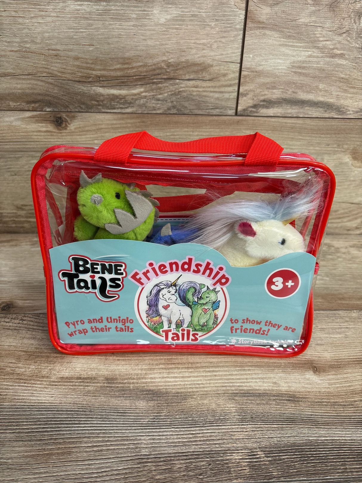 Front view of BeneTails Friendship Tails plush set showcasing green dragon Pyro and white unicorn Uniglo in clear red carry bag on wooden background
