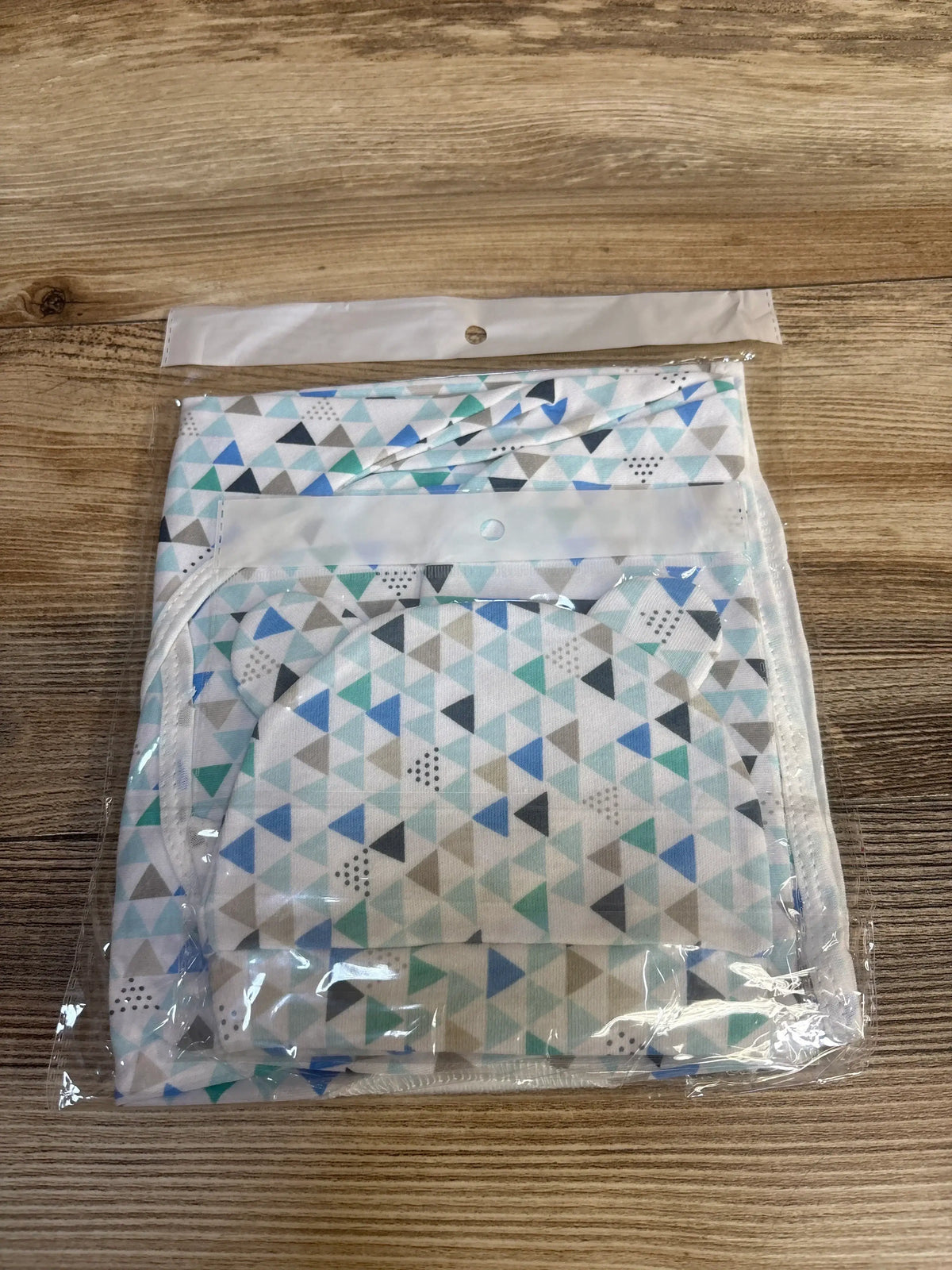 Top-down view of newborn swaddle wrap and matching hat showing blue, teal and gray triangle pattern in clear packaging on wooden surface
