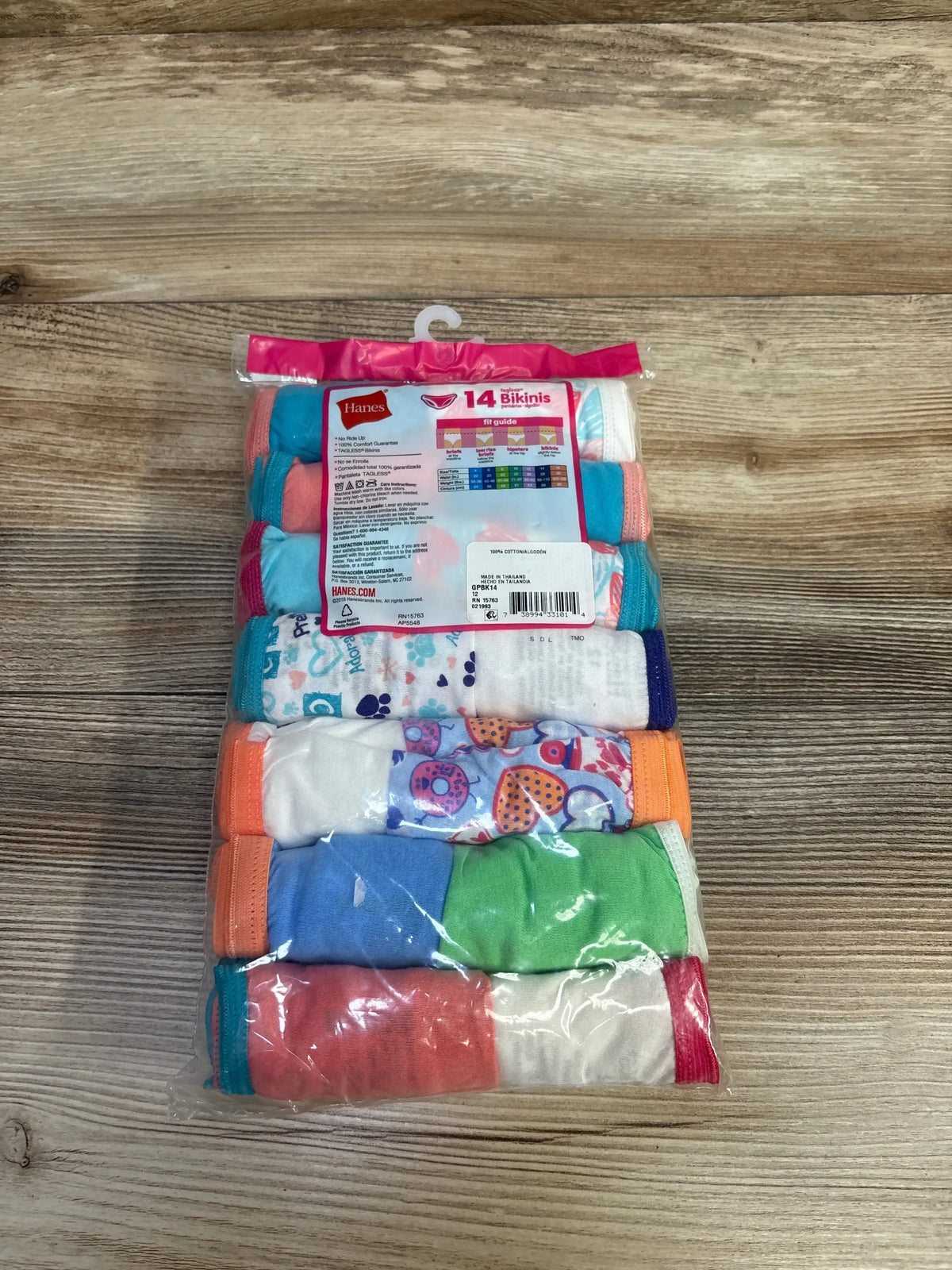 Top-down view of Hanes 14-pack girls’ bikini underwear showing assorted colorful and patterned pairs in clear plastic packaging on wood surface