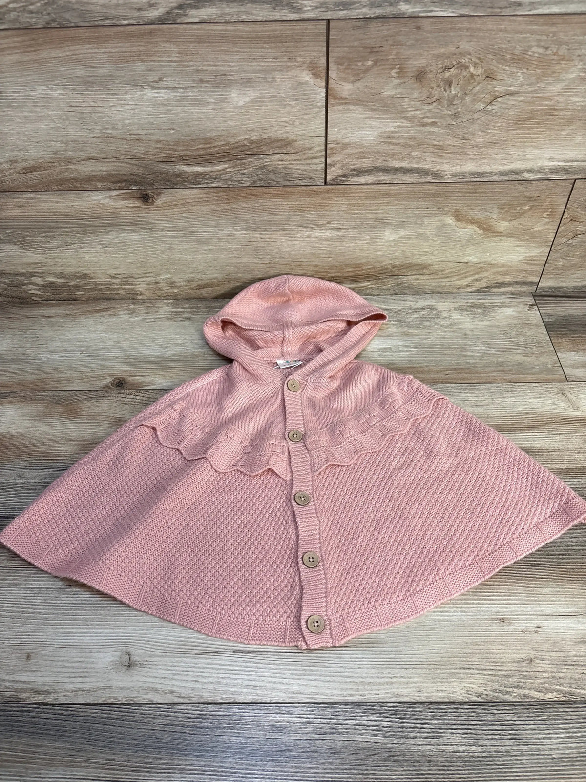 Pink knit hooded toddler cape with buttons, displayed on wood floor