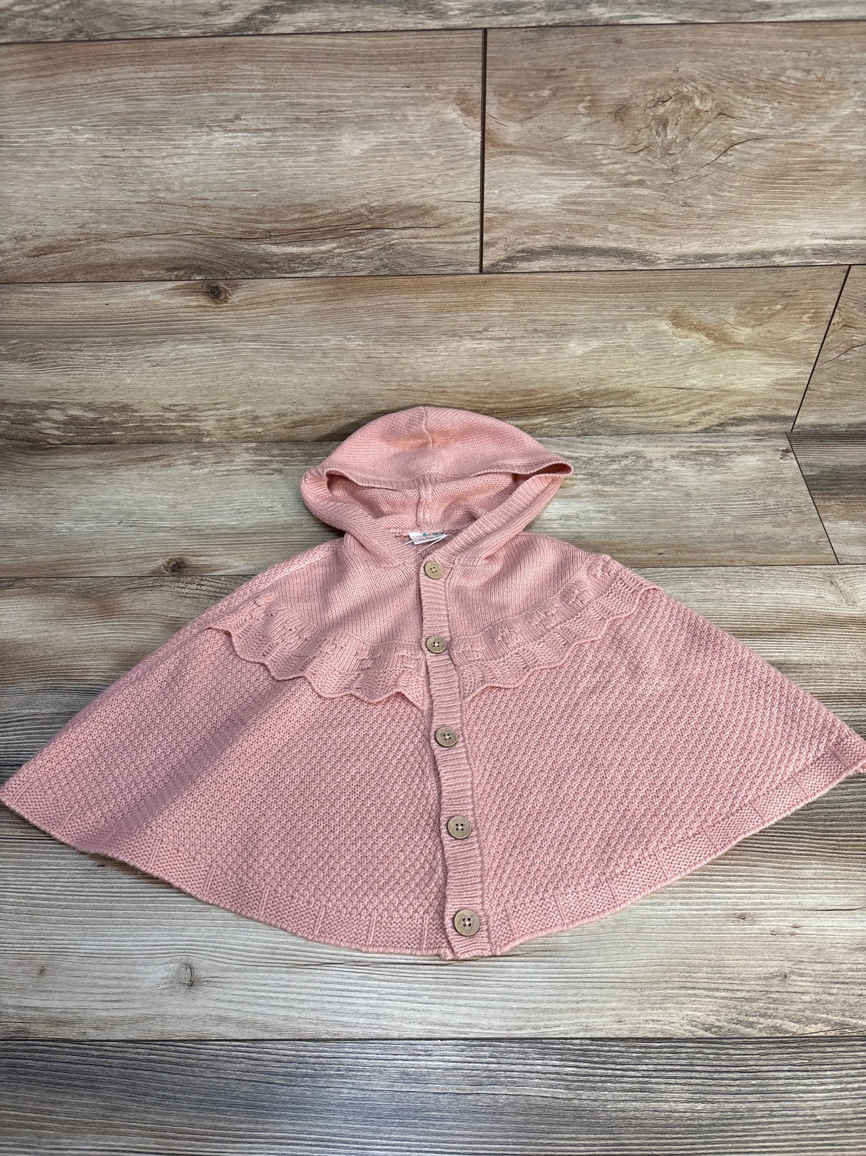 Pink knit hooded toddler cape with buttons, displayed on wood floor