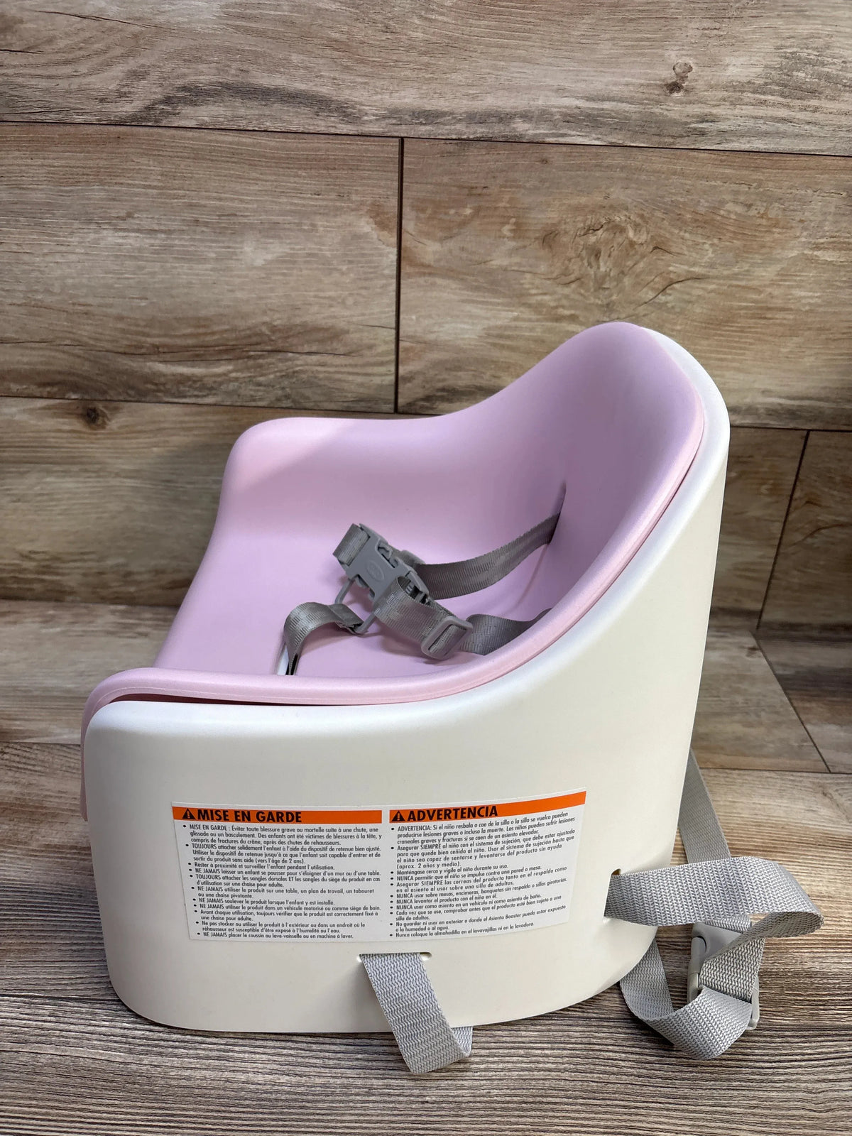 Three-quarter view of OXO Tot Nest booster seat showing blossom pink insert and attached gray safety straps on wood background