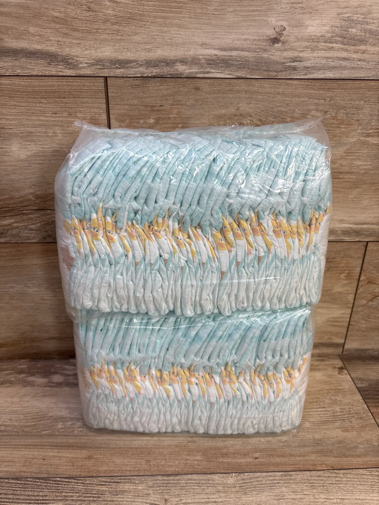 Front view of Pampers Size 1 diapers, two stacked clear-wrapped sleeves showing folded diapers with yellow giraffe print on a wood background