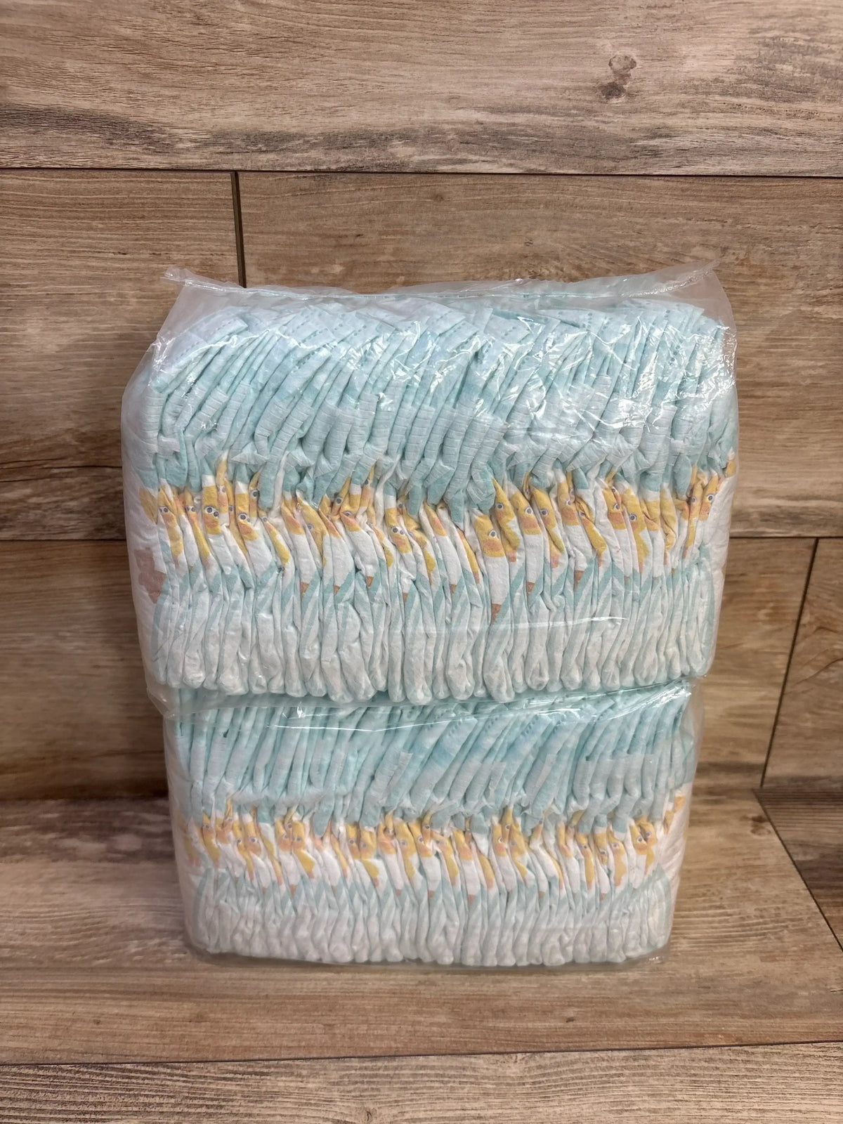 Front view of Pampers Size 1 diapers, two stacked clear-wrapped sleeves showing folded diapers with yellow giraffe print on a wood background