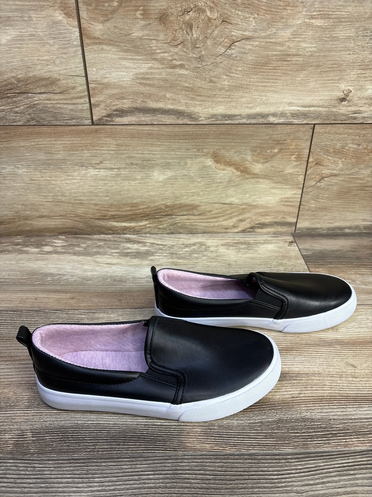 Side view of Cat and Jack Hailey kids’ black slip-on sneakers showing white rubber soles and pink inner lining on wood floor