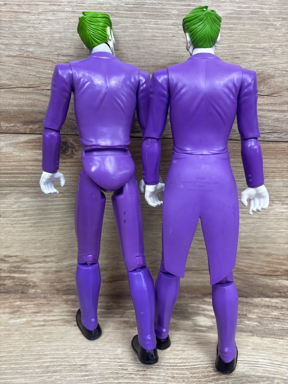 Rear view of two 12-inch DC Comics Joker action figures showcasing purple suits and green hair on a wood-grain background