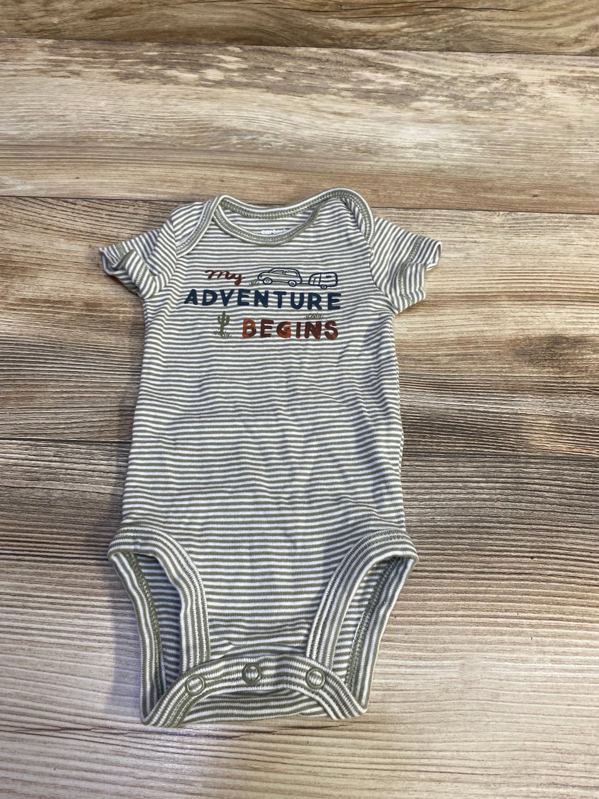 Top-down view of newborn striped green-and-white bodysuit showing My Adventure Begins text and car graphic on wood