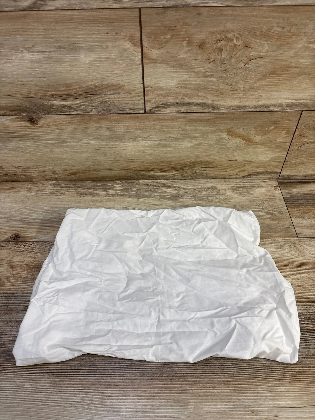 Top-down view of white fitted bassinet sheet with elastic corners and light wrinkles on wood plank background