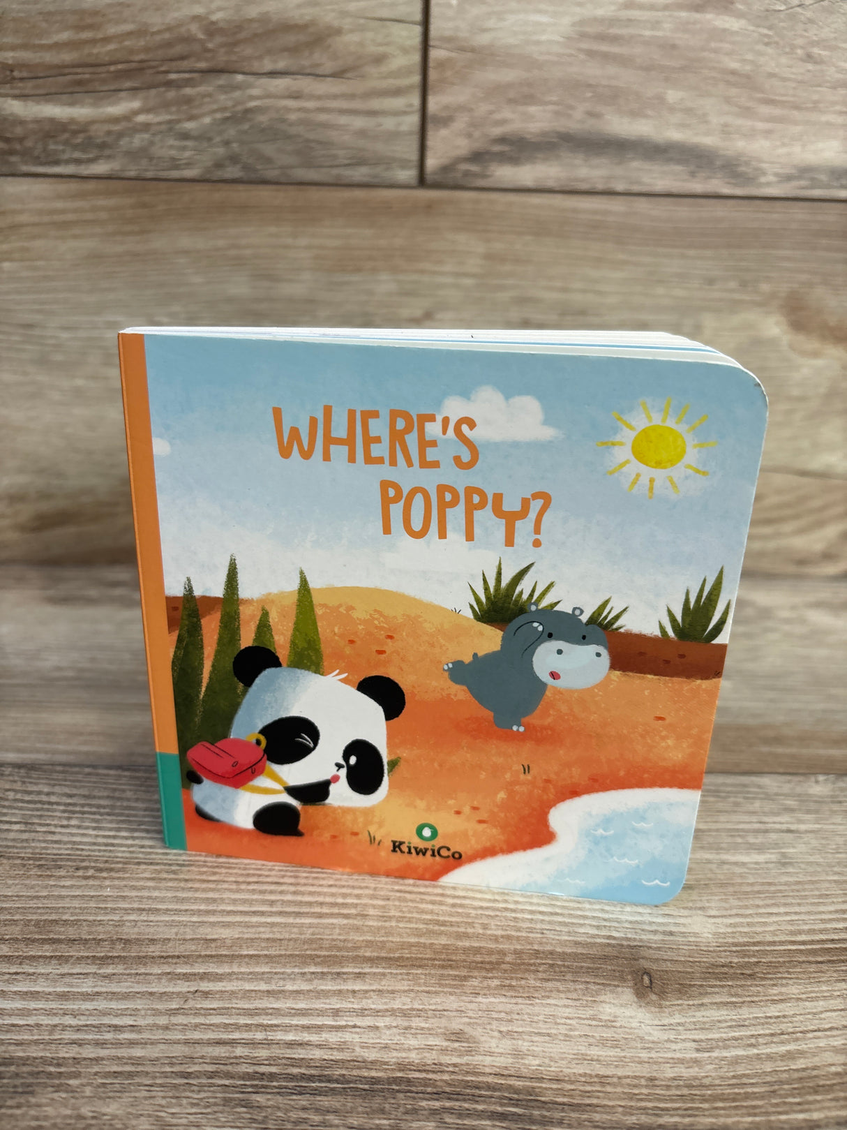 Front cover of KiwiCo Where's Poppy board book showing panda with red bag and hippo on sandy beach, wood tabletop background
