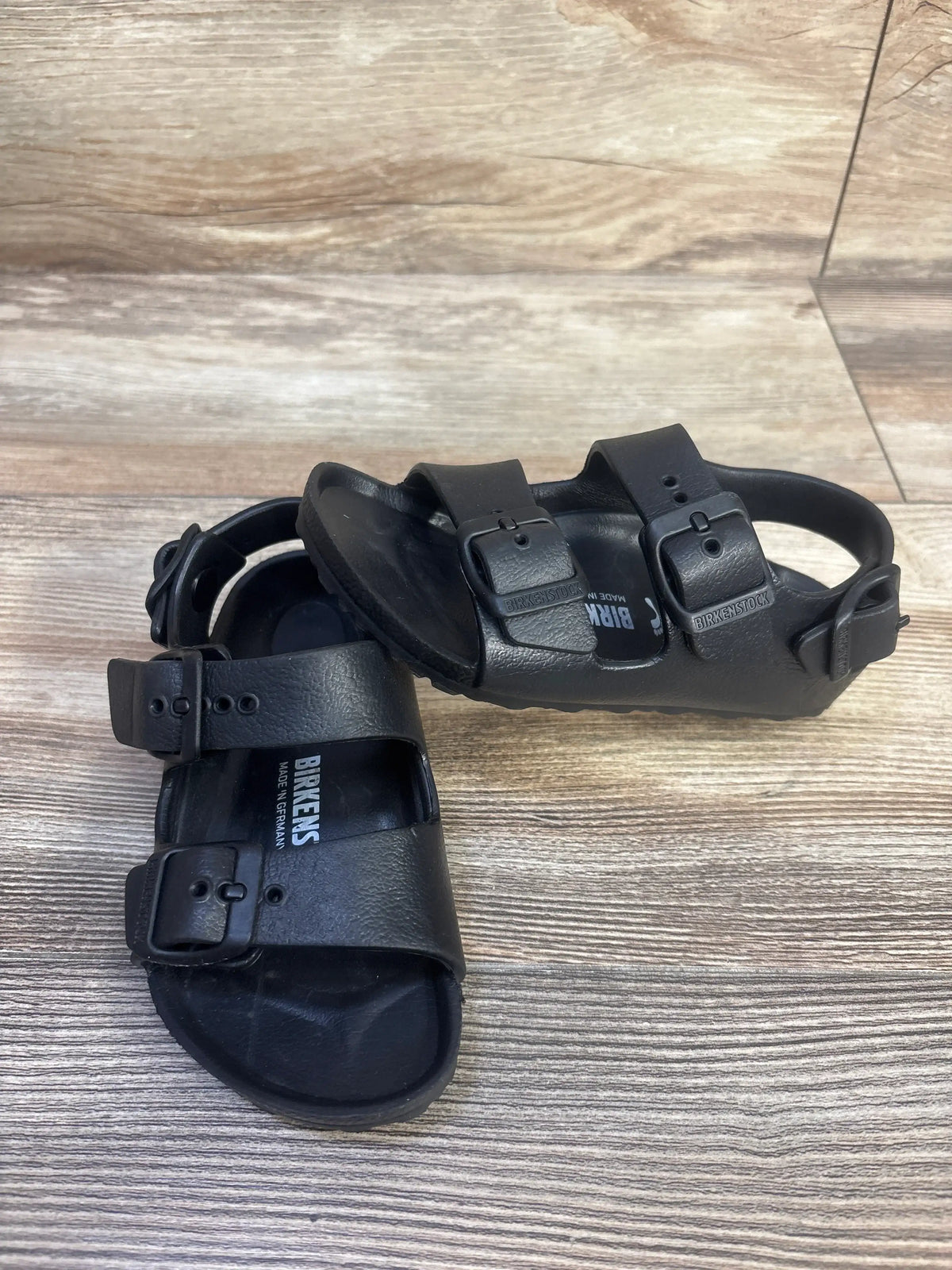 Top view of black Birkenstock Milano EVA sandals showcasing three adjustable straps and backstraps on a wood-grain floor