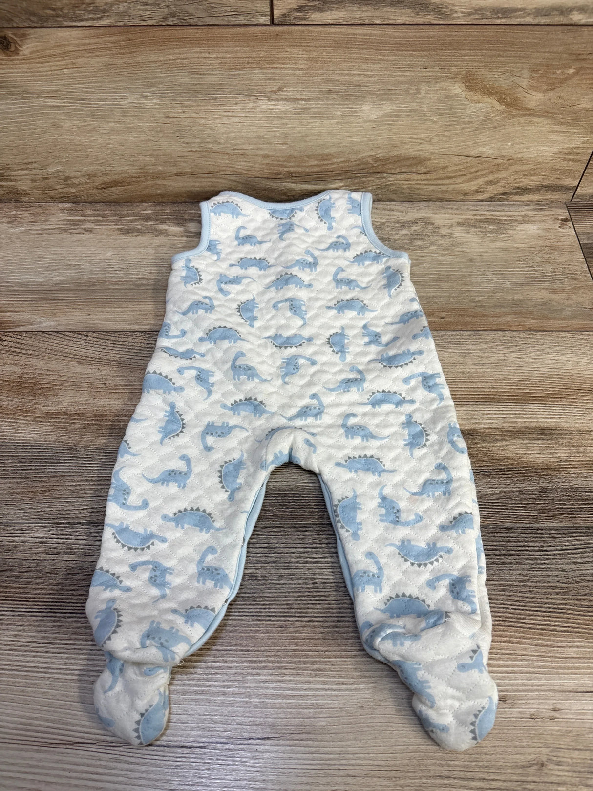Flat lay of Minibean white footed jumpsuit showing blue dinosaur print and quilted texture on wood background, size 6-9 months
