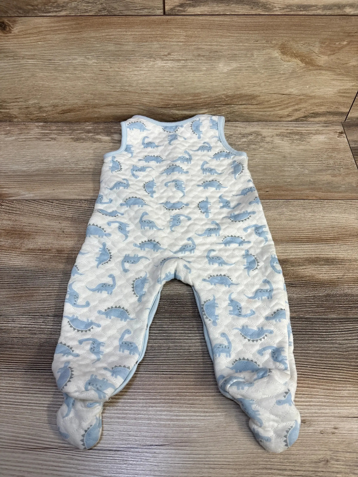 Flat lay of Minibean sleeveless footed jumpsuit in white with blue dinosaur print and quilted texture on wood plank floor