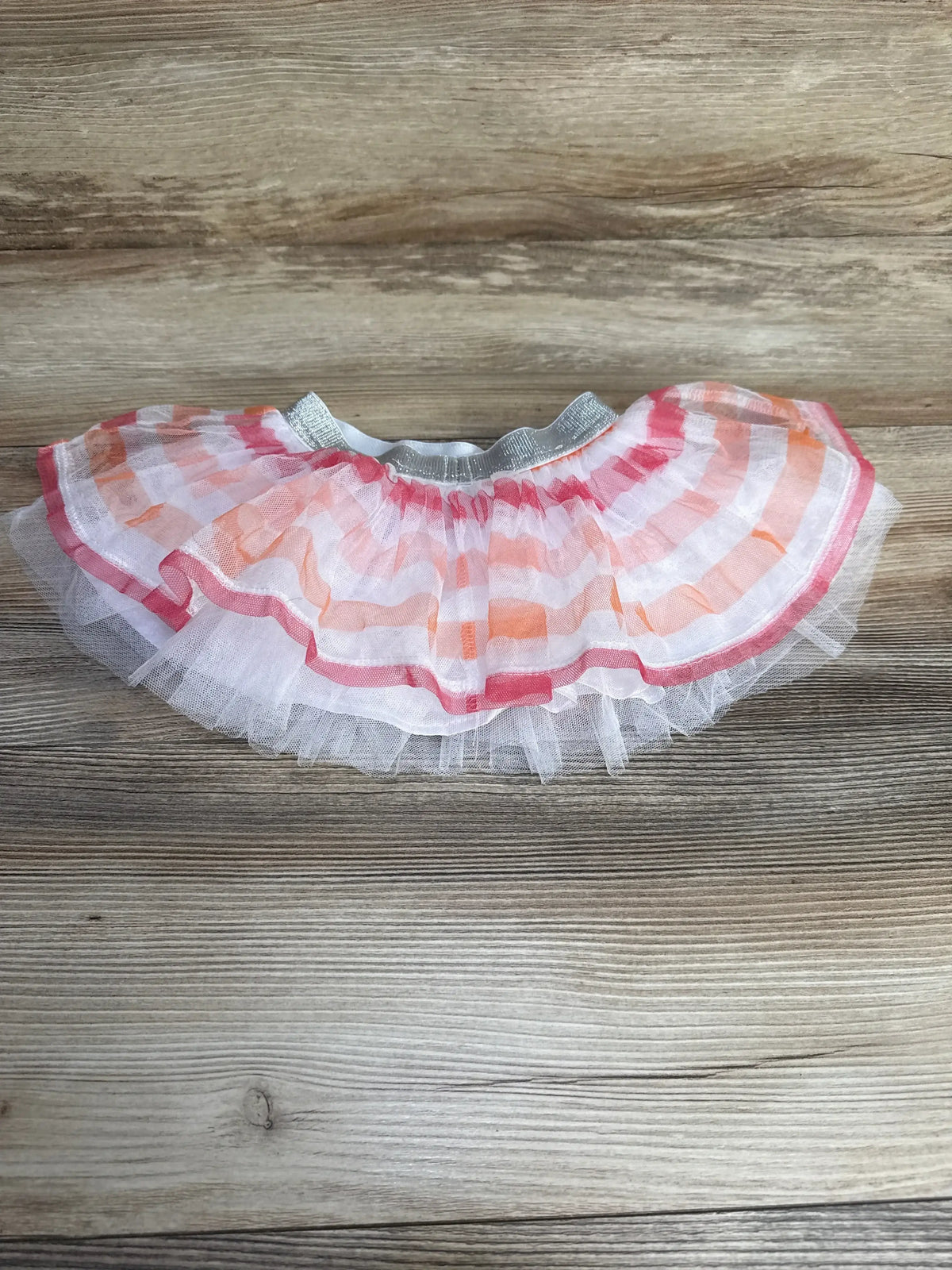Flat lay of baby tutu skirt with silver elastic waistband, white tulle and pink and orange striped overlay on wood background