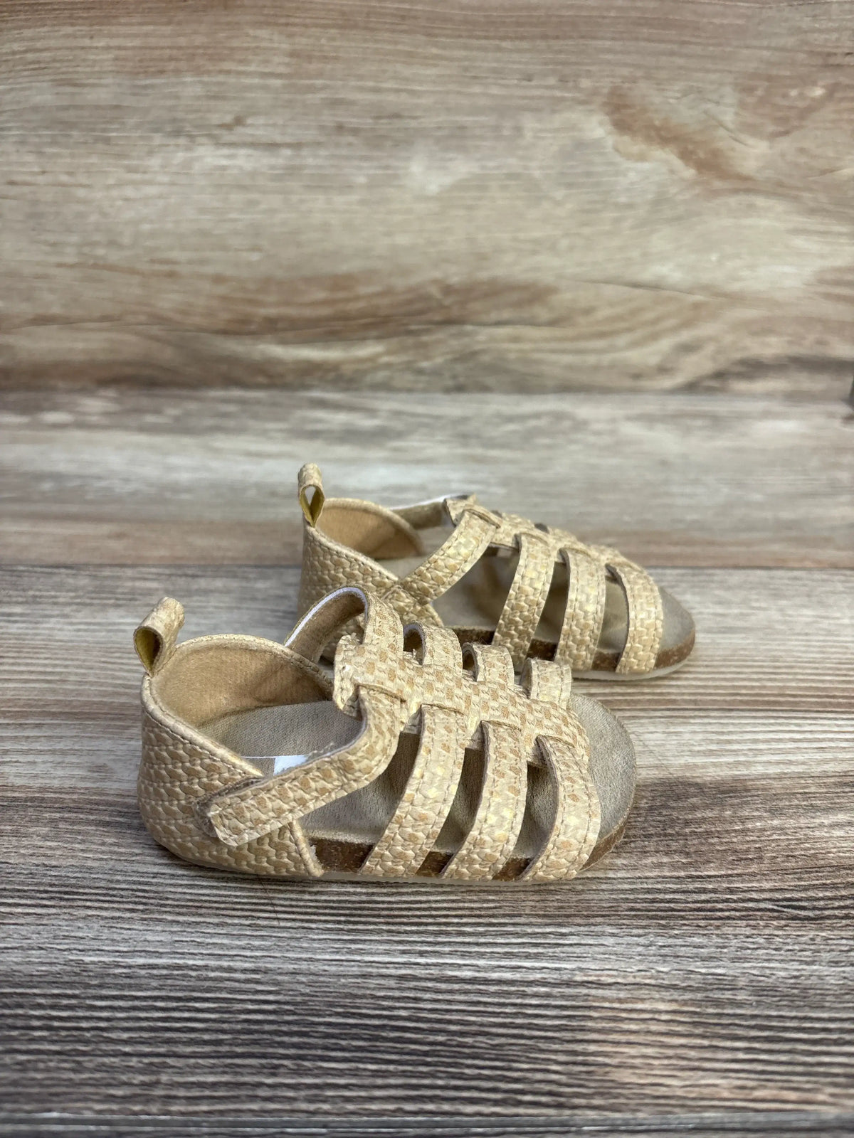 Three-quarter view of Carter’s strappy sandals in solid brown with textured straps and ankle loops on wood-grain background