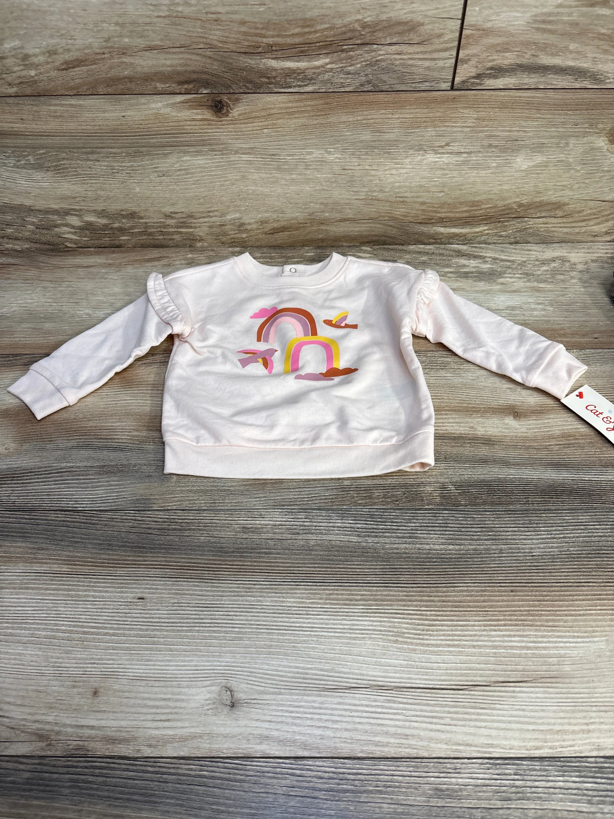 Front flat lay of pink Cat & Jack baby sweatshirt showcasing rainbow and cloud graphic and ruffled shoulder detail on wood background