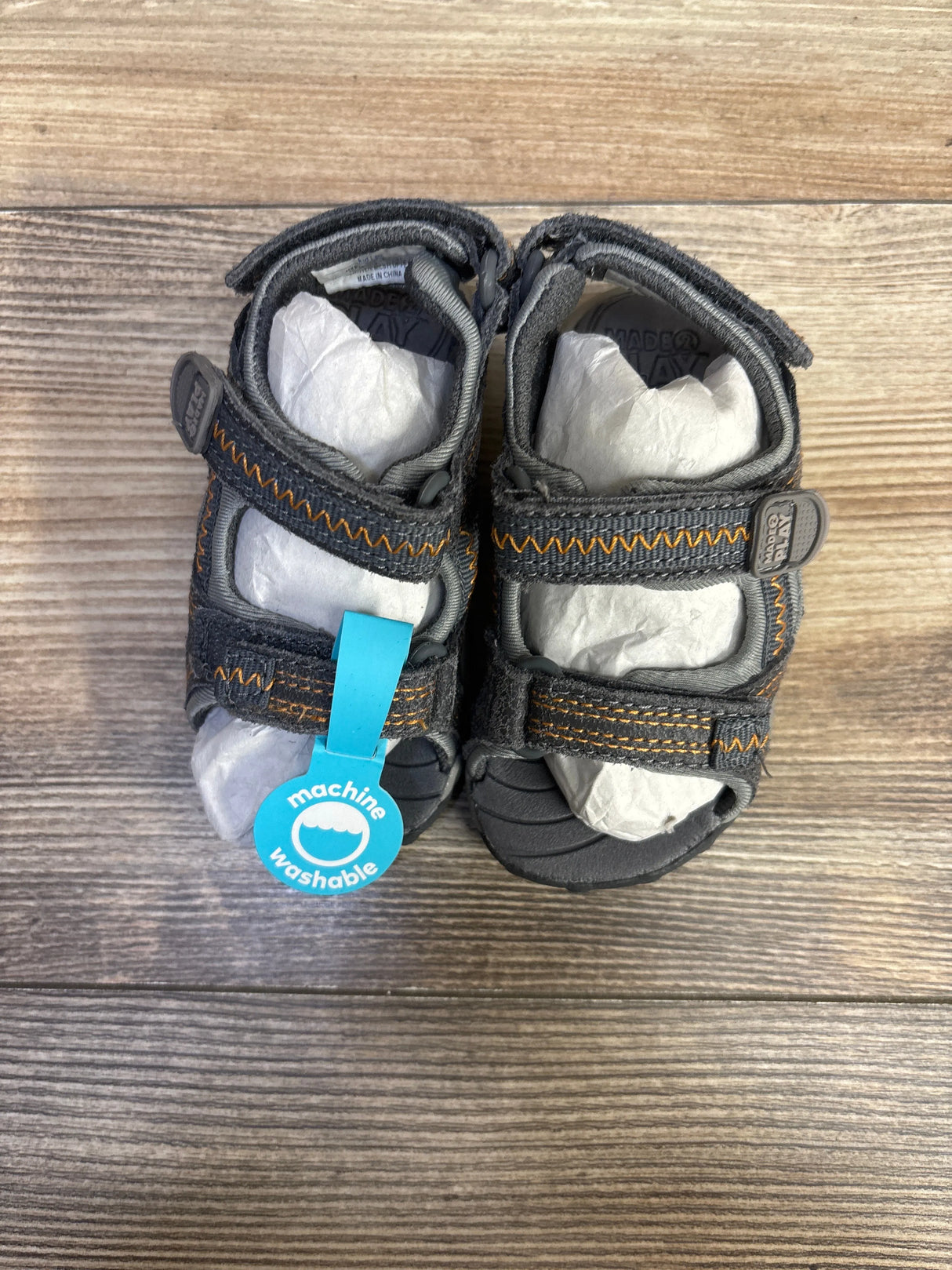 Top-down view of Stride Rite Made 2 Play Issac toddler sandals showing grey fabric with orange stitching, adjustable Velcro straps, and rubber toe bumper on wood floor
