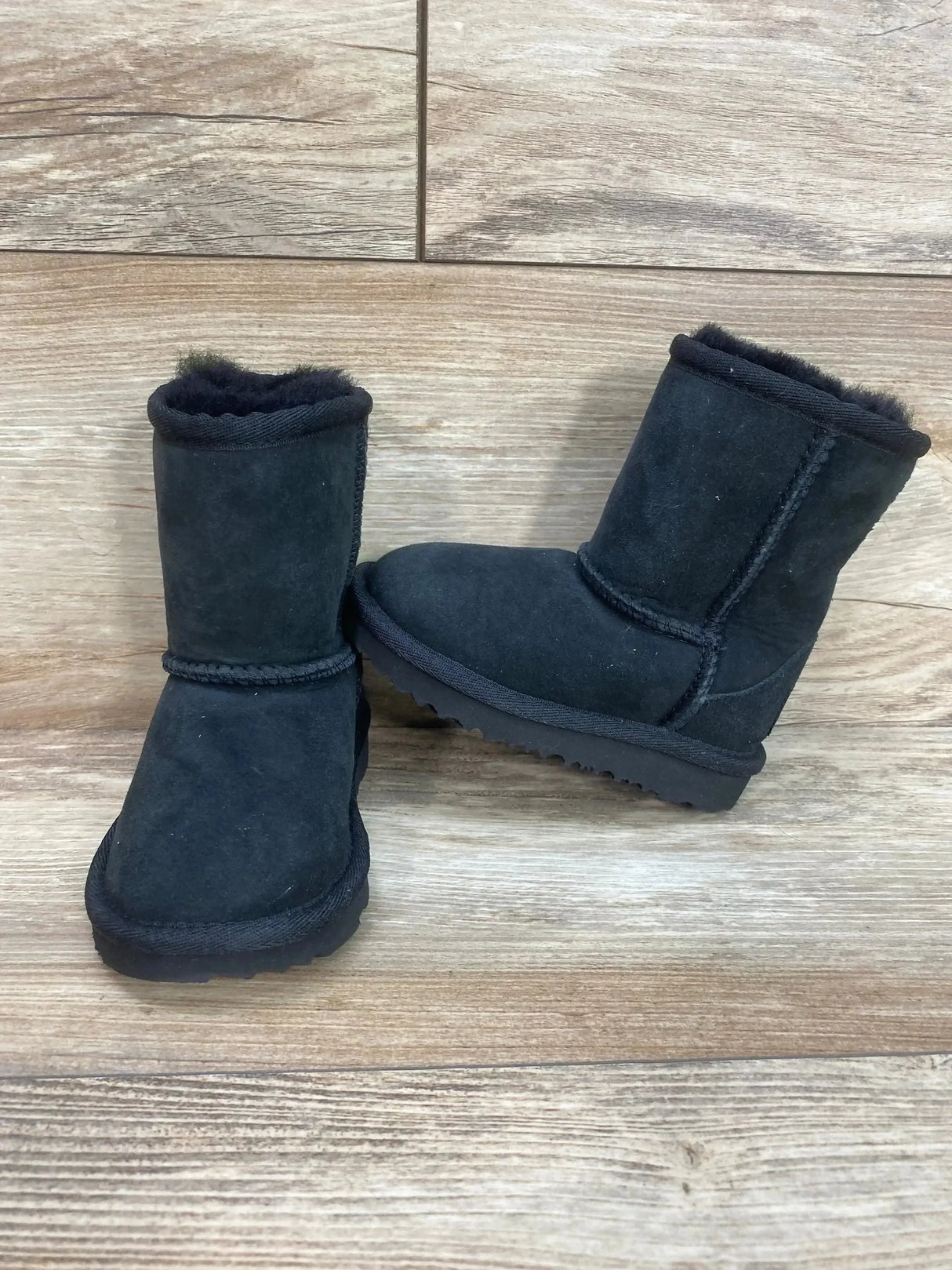 Three-quarter view of UGG Toddlers Classic II Boot in black showcasing suede uppers with fleece lining on wood plank floor