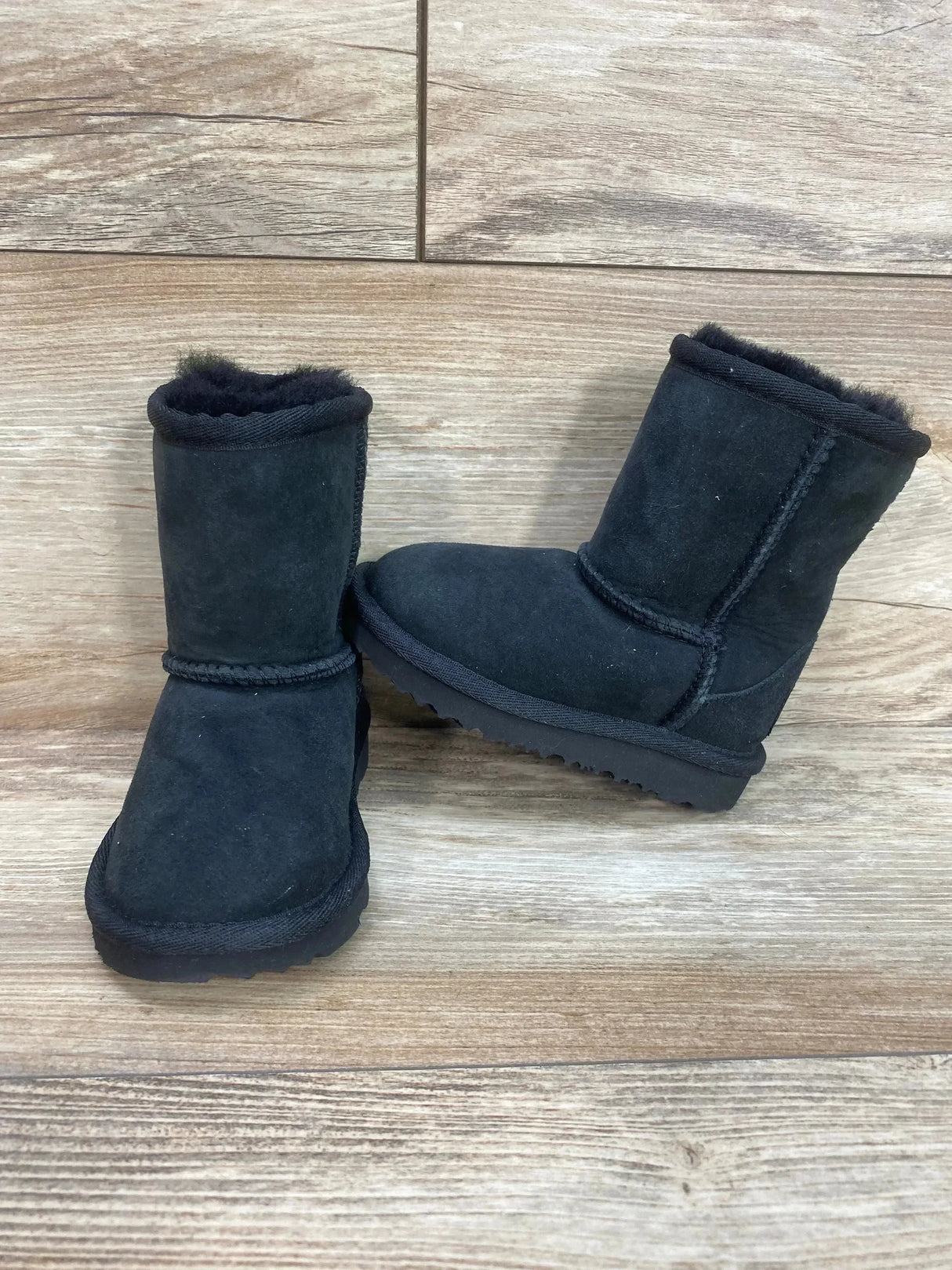 Front and side view of UGG toddler Classic II black boot with suede upper, plush lining and textured sole on wood floor