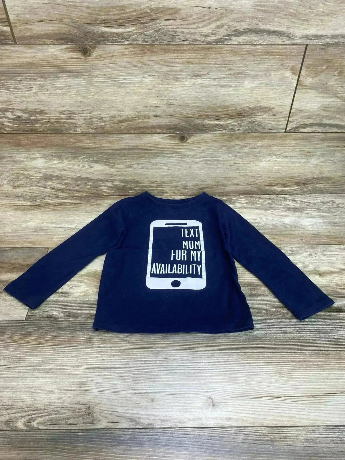 Flat lay of Children’s Place Text Mom For My Availability Shirt size 4T showcasing navy long-sleeve tee with phone graphic print on wood plank floor